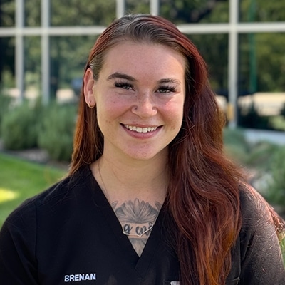A woman with long reddish-brown hair, wearing black medical scrubs with the name "Brenan" embroidered, smiles outside in front of a building with large windows and greenery in the background.