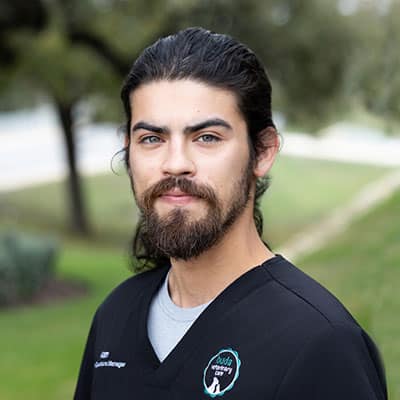 A man with long dark hair and a beard, wearing a black scrub top with a veterinary clinic logo, stands outside on a grassy area with trees in the background, looking at the camera.