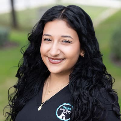 Smiling woman with long, curly black hair wearing a black shirt with a "buda veterinary care" logo, standing outdoors with greenery in the blurred background.