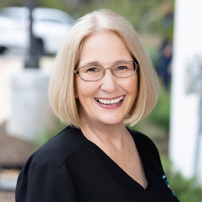 A smiling woman with straight, shoulder-length blonde hair and glasses wears a black top and stands outdoors with a blurred background of greenery and a parked car.