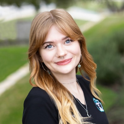 A young woman with long, wavy, light brown hair and blue eyes smiles outdoors. She wears a black top, hoop earrings, layered necklaces, and has a nose ring. The background is grassy and blurred.