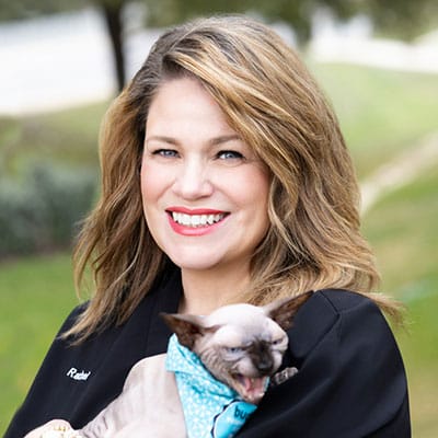 A smiling woman with wavy, shoulder-length hair holds a hairless cat wearing a blue bandana. The cat has its mouth open, showing teeth. They are outdoors with greenery in the background.