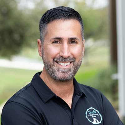 Smiling man with short dark hair and a beard, wearing a black polo shirt with a "buda veterinary clinic" logo, standing outdoors with blurred greenery in the background.