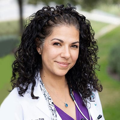 A woman with curly dark hair wearing a white medical coat and a purple top, smiling outdoors with greenery in the background. She has a stethoscope around her neck.