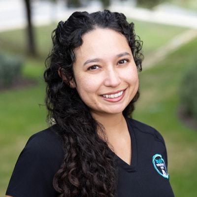 A woman with long, curly dark hair smiles outdoors. She is wearing a black shirt with a blue and white logo. The background is blurred greenery and a walkway.