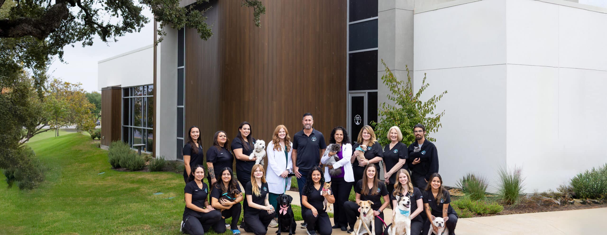 A group of veterinary staff in black uniforms pose with several dogs and cats outside a modern animal clinic building on a grassy lawn.
