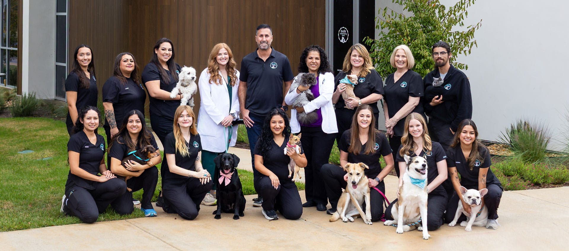 A group of 16 smiling people, some holding or sitting beside dogs of various breeds and sizes, pose in front of a building with greenery and a sidewalk. Most wear matching black outfits; two wear white coats.