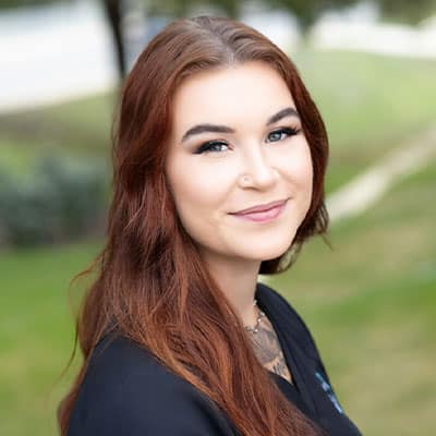 A young woman with long reddish-brown hair and subtle makeup smiles while standing outdoors, with a blurred green grassy background. She is wearing a black top.