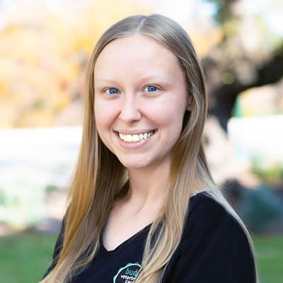 A young woman with long straight blonde hair smiles at the camera outdoors. She is wearing a black top and the background is softly blurred with greenery and sunlight.