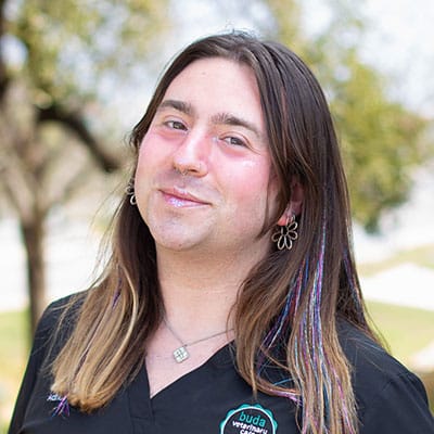 A person with long brown hair, wearing a black top with a logo, flower-shaped earrings, and a necklace, stands outdoors with trees and a bright sky in the background, smiling gently at the camera.