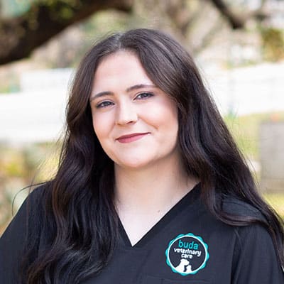 A woman with long dark hair wearing a black scrub top with a "buda veterinary care" logo stands outdoors, smiling gently at the camera. The background is blurred with trees and greenery.