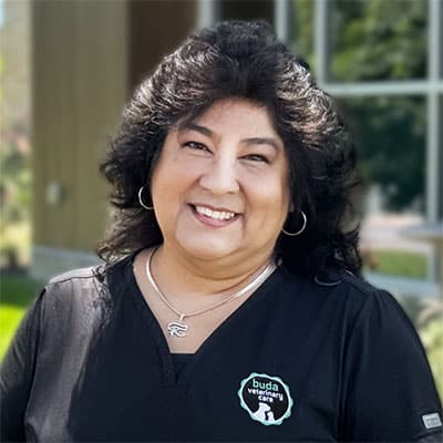 Smiling woman with dark, wavy hair wearing a black scrub top with a "buda veterinary" logo, standing outdoors in front of a building and greenery.