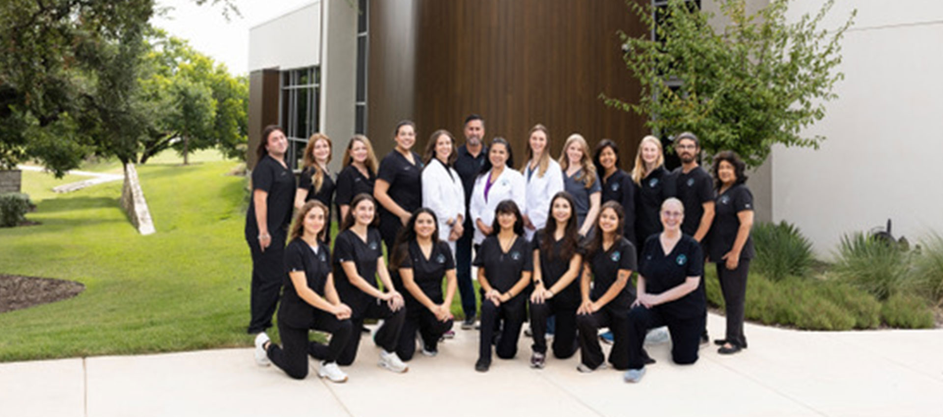 A group of 20 adults, some in black uniforms and others in white coats, pose together outdoors in front of a modern building with trees and grass in the background.