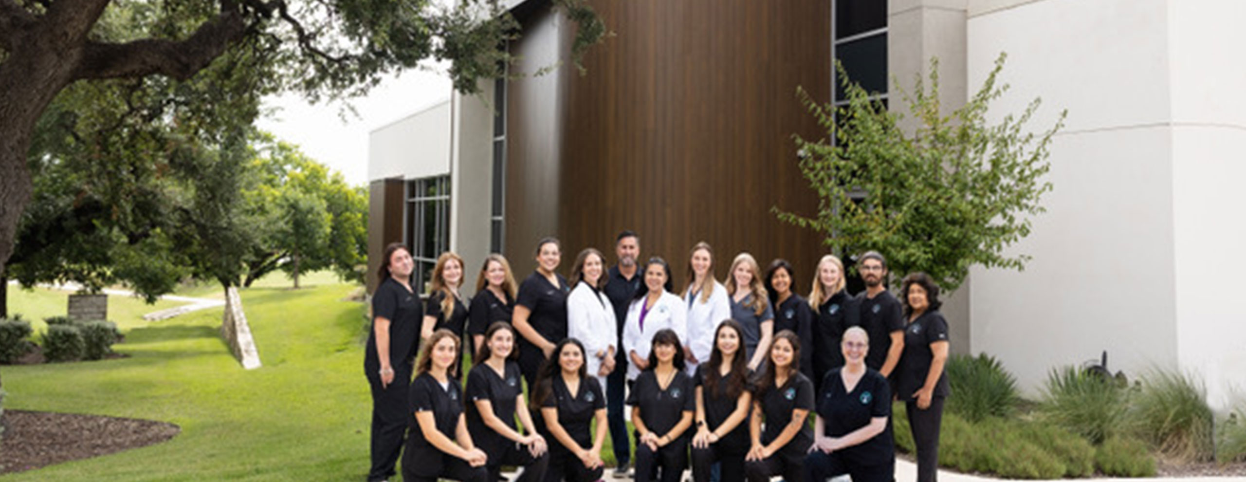 A group of people in matching black uniforms and a few in white coats pose together outside a modern building surrounded by trees and grass.