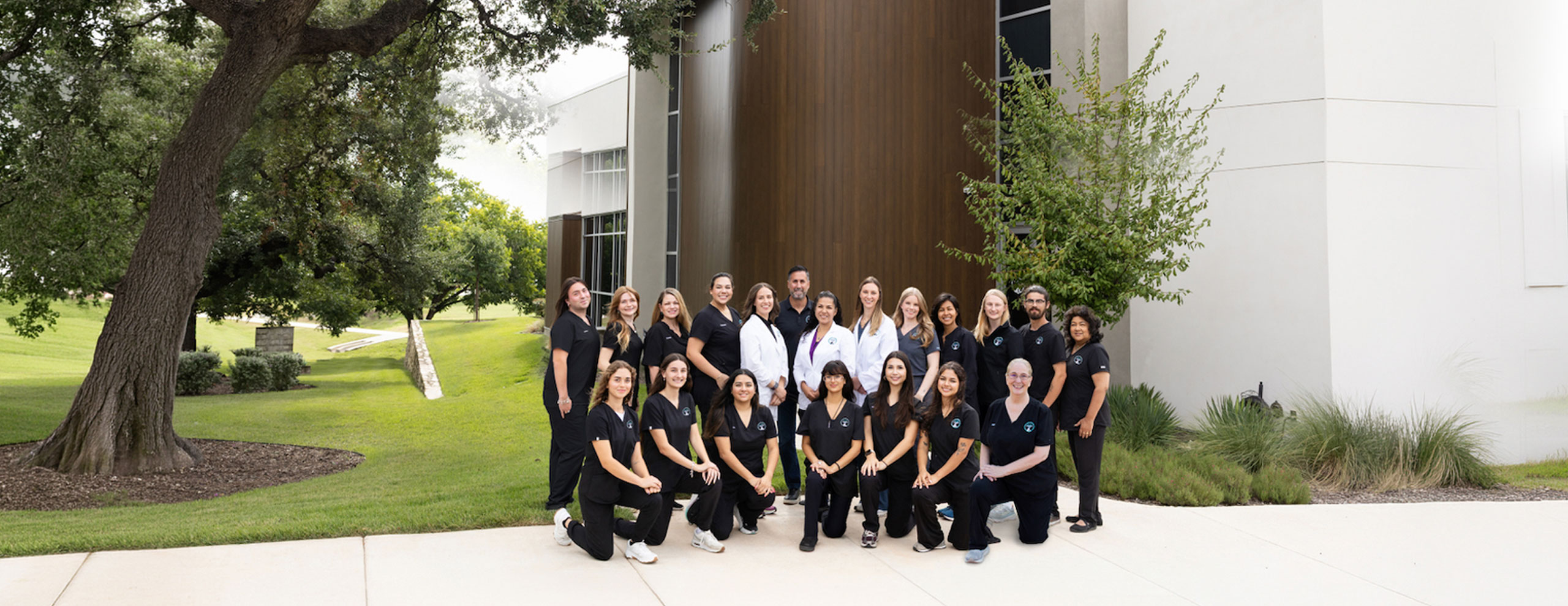 A group of 22 people, mostly women, pose outside a modern building with trees and grass, all wearing matching black uniforms; two people in the center wear white coats.